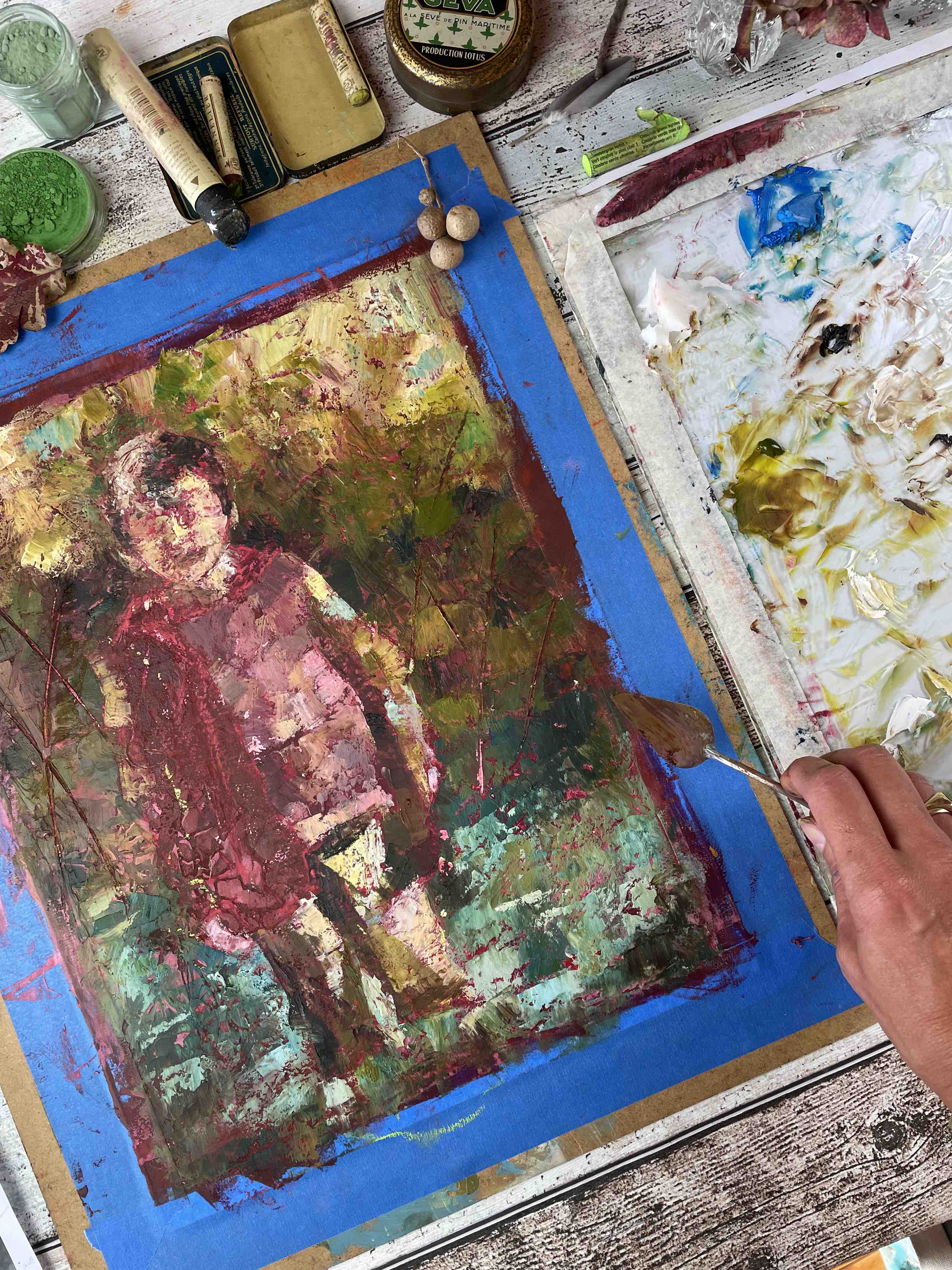 Marina ajoute de la peinture au Ramasseur de brindilles avec un couteau à peindre, entourée de couleurs et d’outils sur sa table d’atelier